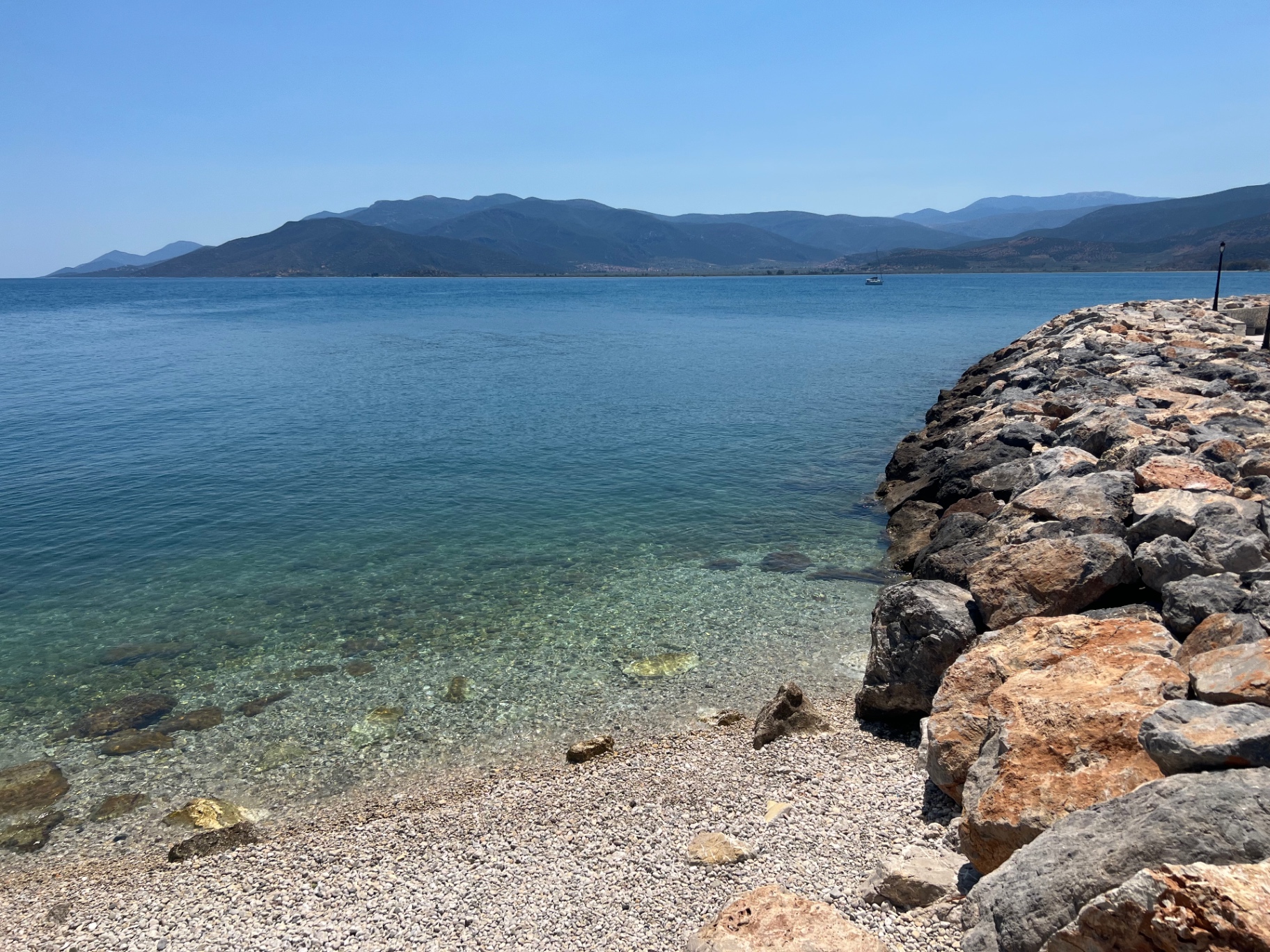 Clear turquoise water and a stone breakwater at the harbour of Paralio Astros, with mountains across the bay