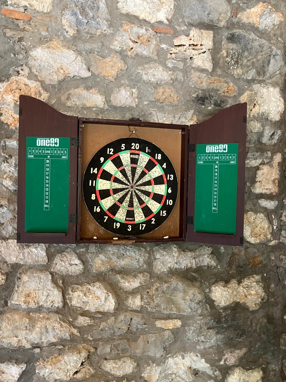 Dartboard mounted in a wooden cabinet on the stone wall of the Villa Arcadia game room