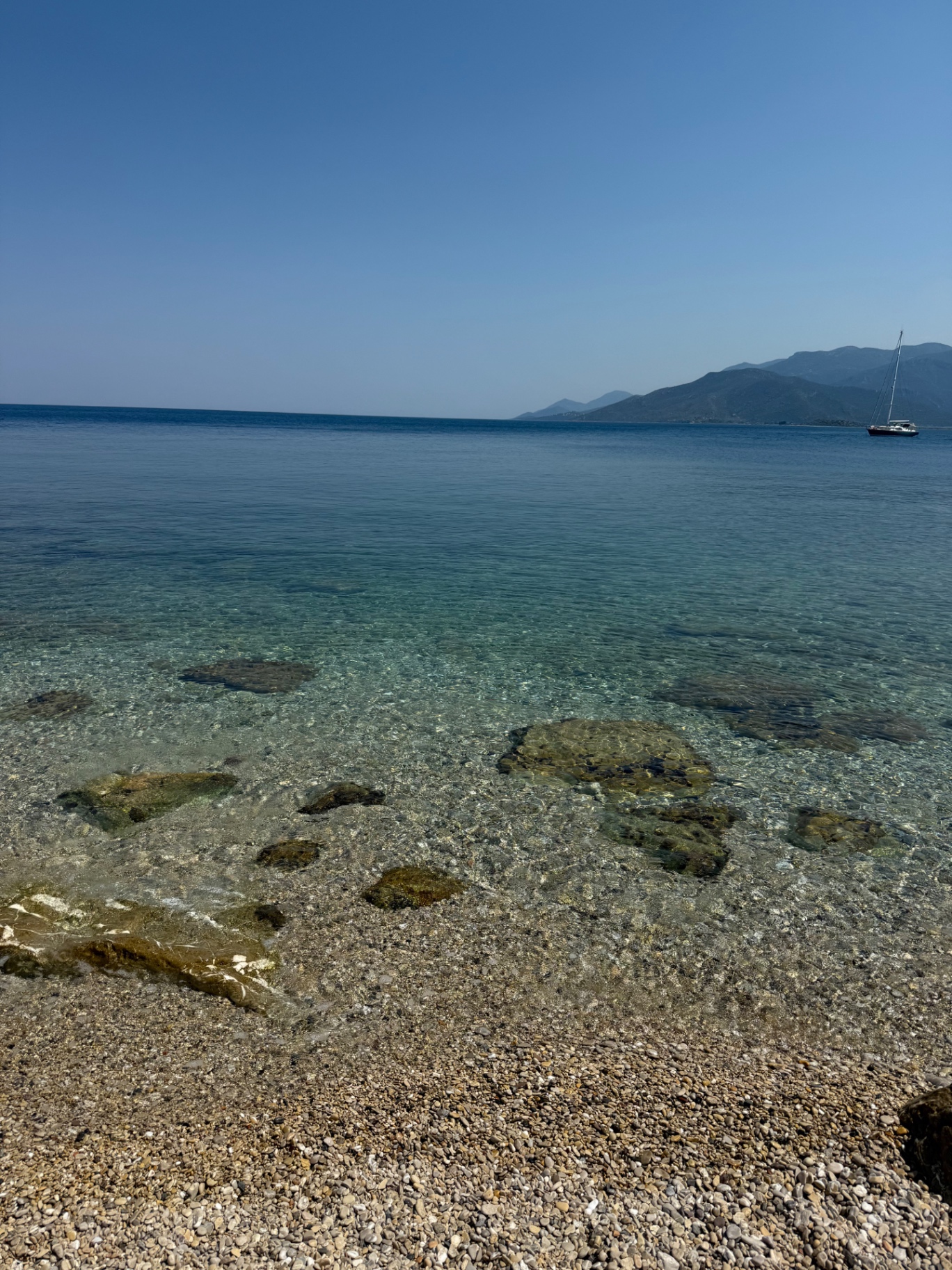 Crystal-clear shallow water over a pebble beach south of Paralio Astros with a sailboat on the horizon